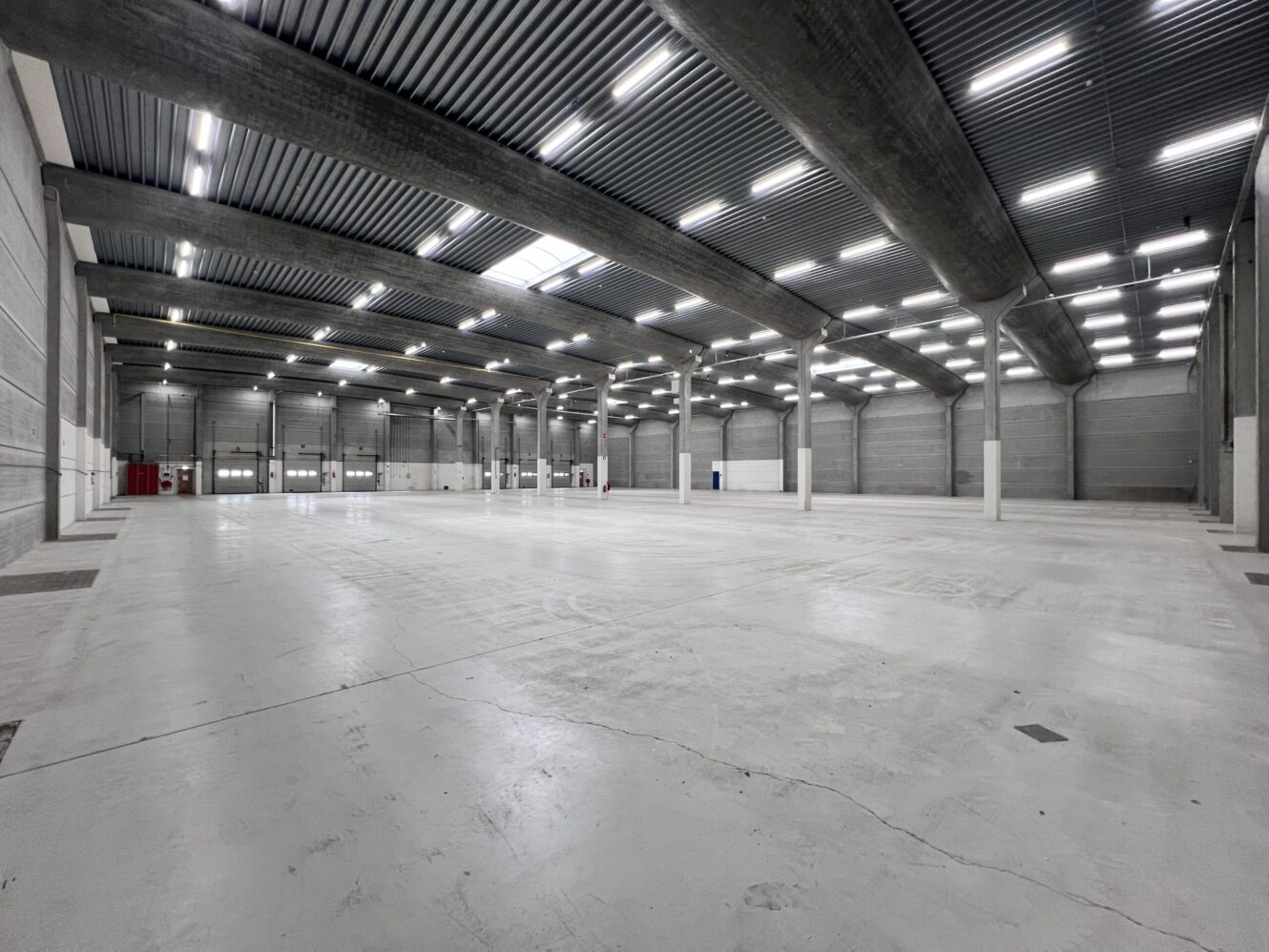 Ruim, leeg industrieel pakhuis met hoge plafonds, betonnen vloeren, grote steunpilaren en overheadverlichting.