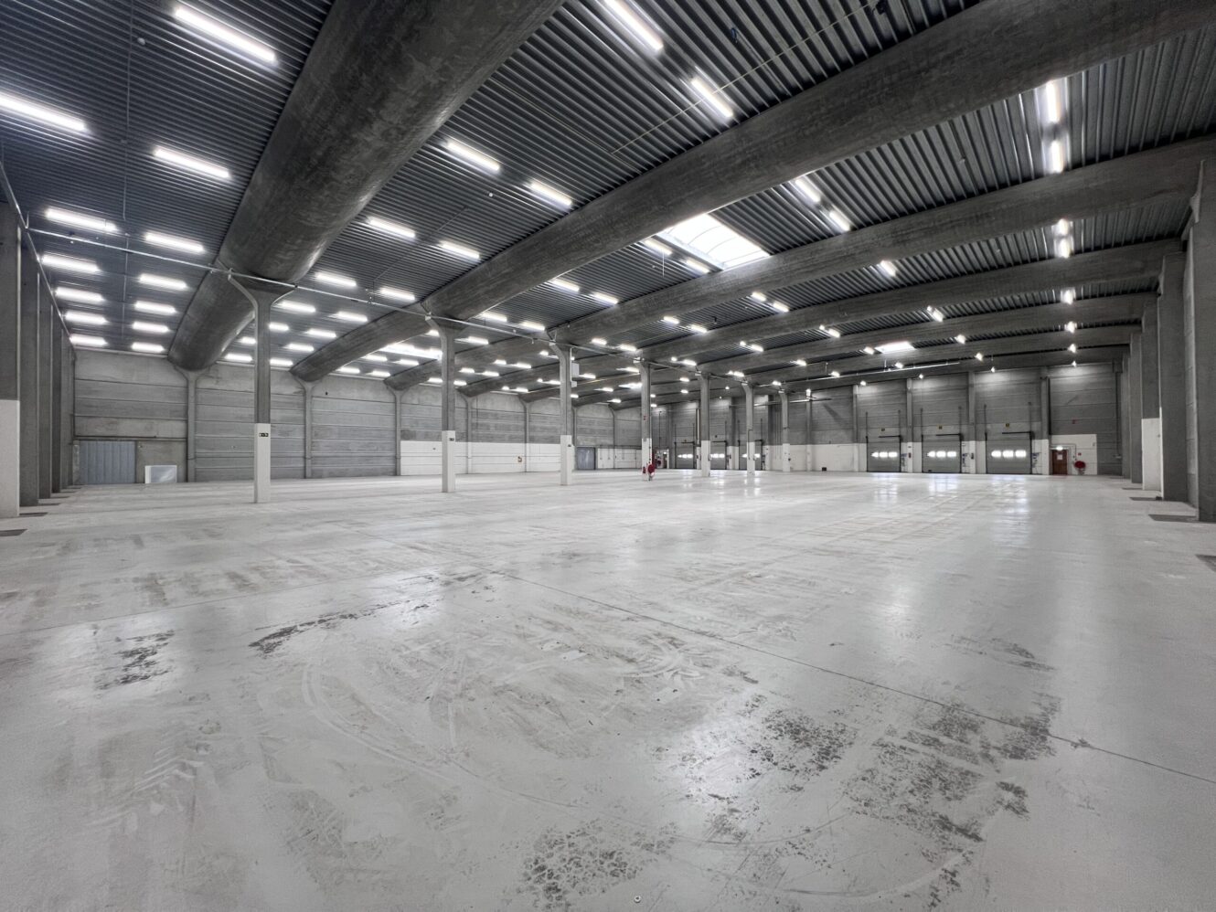 Groot, leeg industrieel pakhuis met betonnen vloeren, hoge plafonds en bovenverlichting. Structurele kolommen en muren omzomen het ruime interieur.