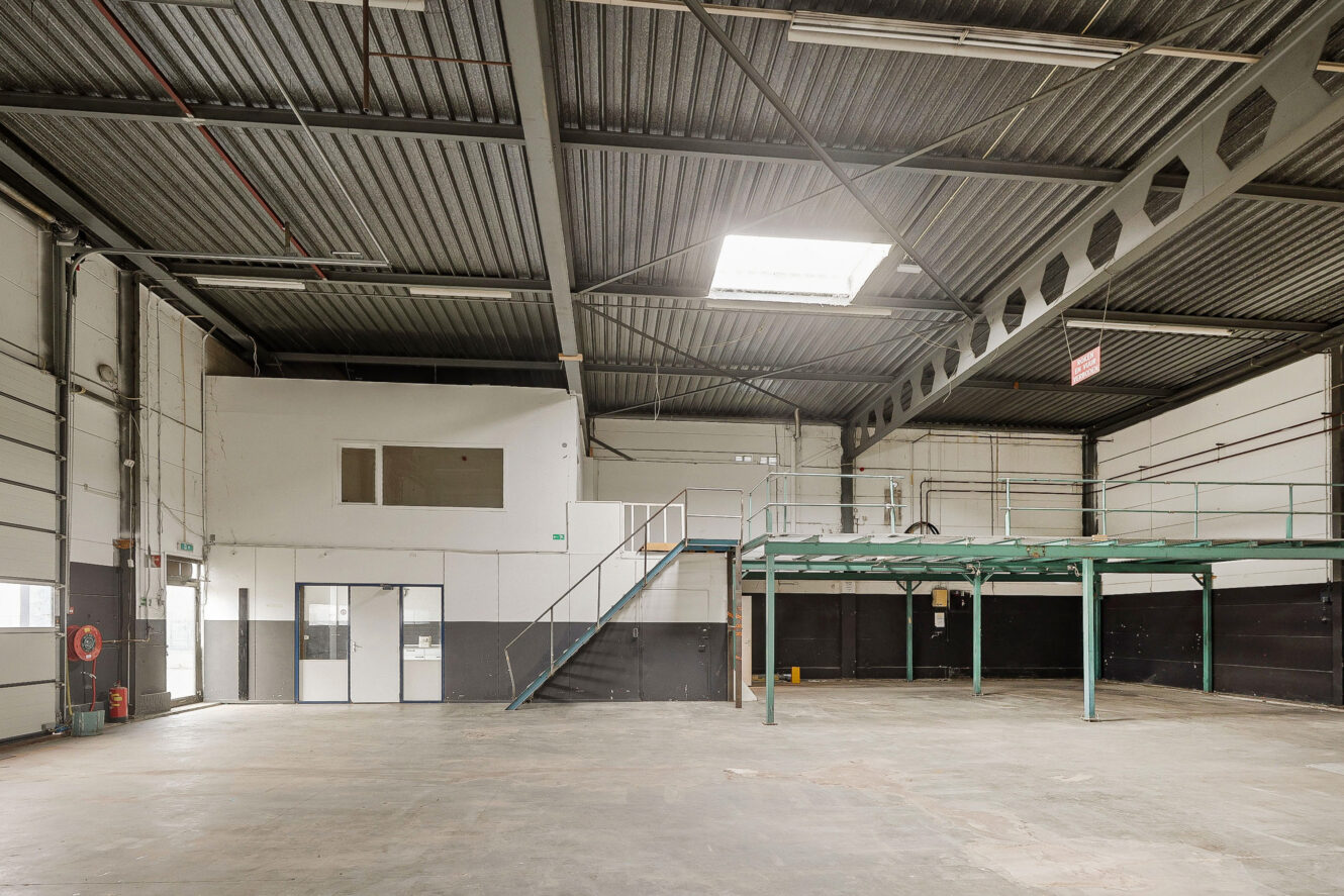 Leeg industrieel magazijninterieur met betonnen vloer, hoog plafond, tussenverdieping en kantoorgedeelte met ramen en deuren aan de linkerkant.