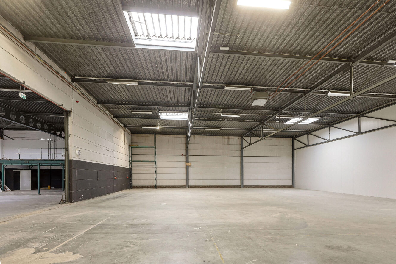 Grote, lege industriële opslagruimte met betonnen vloer, hoog metalen golfplaten plafond, dakramen en witte paneelmuren.