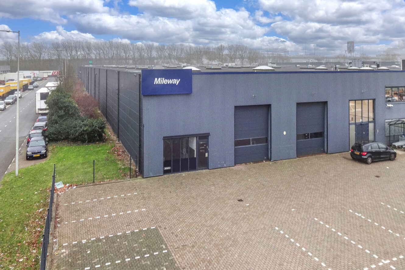 Een groot, modern magazijngebouw met een Mileway-bord, parkeerplaatsen aan de voorkant en een paar auto's geparkeerd in de buurt onder een gedeeltelijk bewolkte hemel.