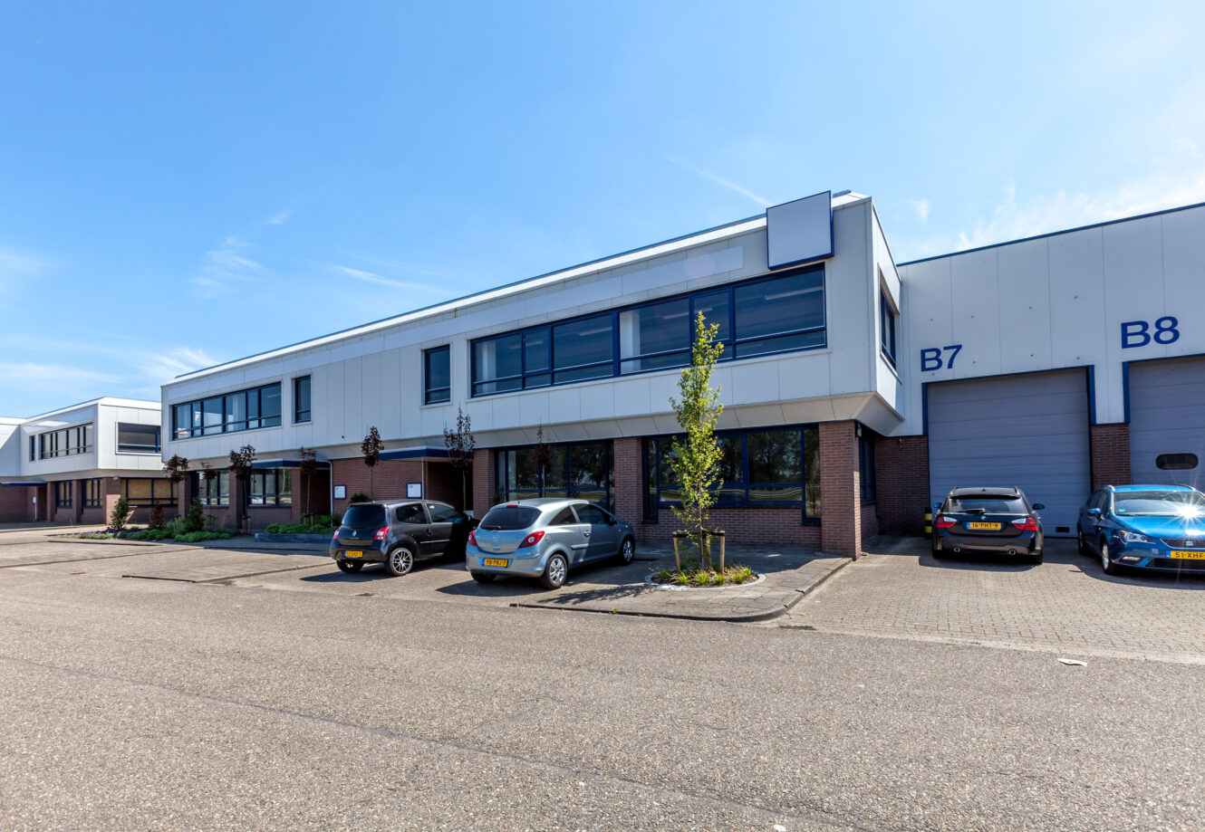 Een modern industrieel gebouw met grote ramen, parkeerplaatsen en geparkeerde auto's aan de voorkant. Het gebouw heeft het label B7 en B8. De lucht is helder en zonnig.