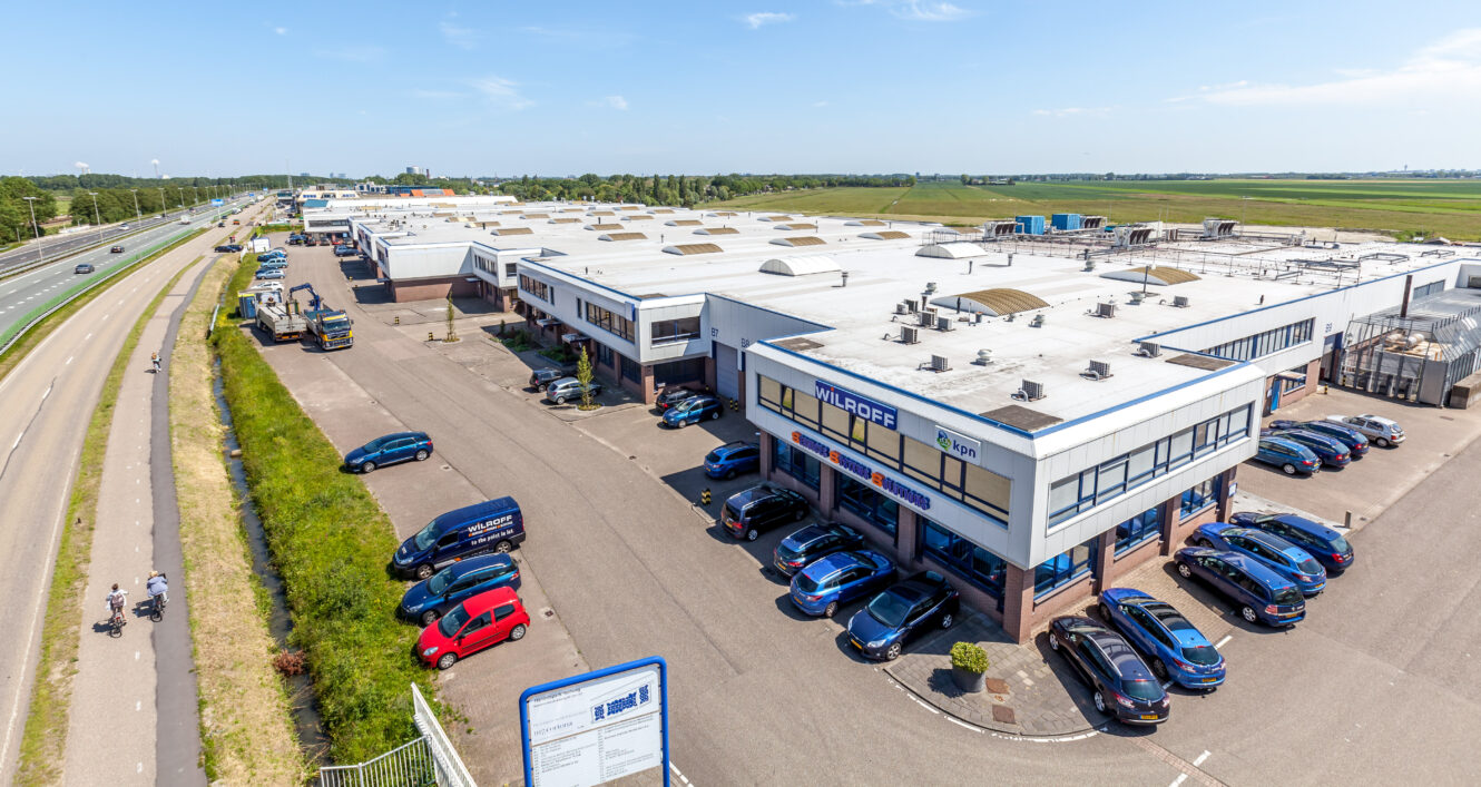 Luchtfoto van een industrieterrein met meerdere magazijnen, geparkeerde auto's en vrachtwagens; op een heldere dag loopt er een weg langs het complex.