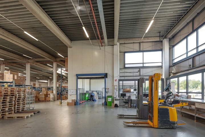 Ruim magazijninterieur met pallets, dozen, rekken en industriële apparatuur, waaronder een gele vorkheftruck, onder hoge plafonds met grote ramen.