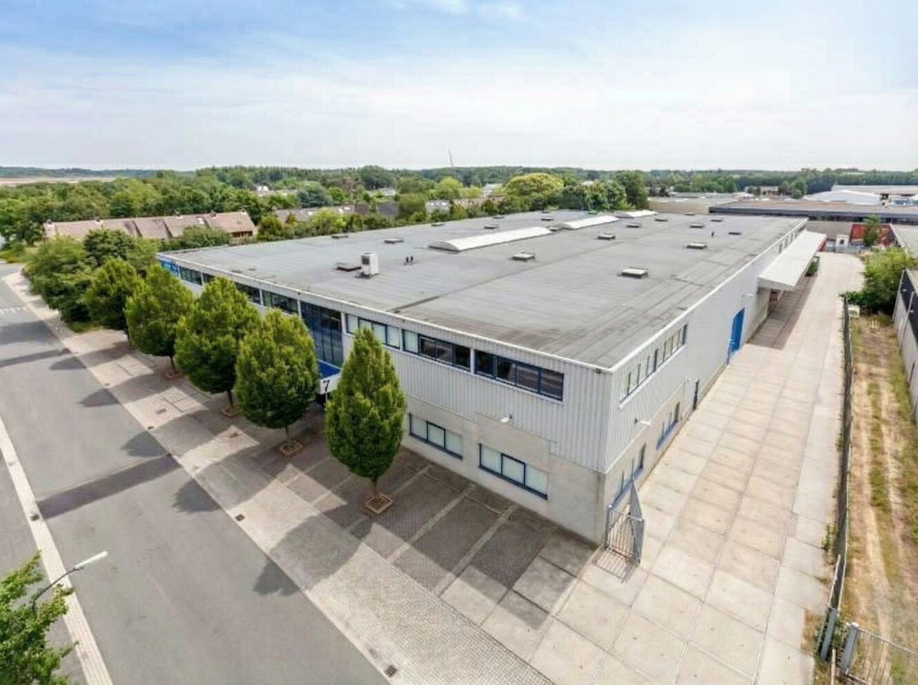 Een groot industrieel pakhuis met een plat dak, omringd door bomen en betonnen bestrating, naast een rustige straat onder een gedeeltelijk bewolkte hemel.