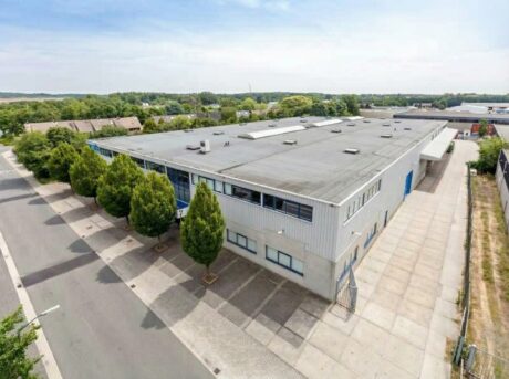Een groot industrieel pakhuis met een plat dak, omringd door bomen en betonnen bestrating, naast een rustige straat onder een gedeeltelijk bewolkte hemel.