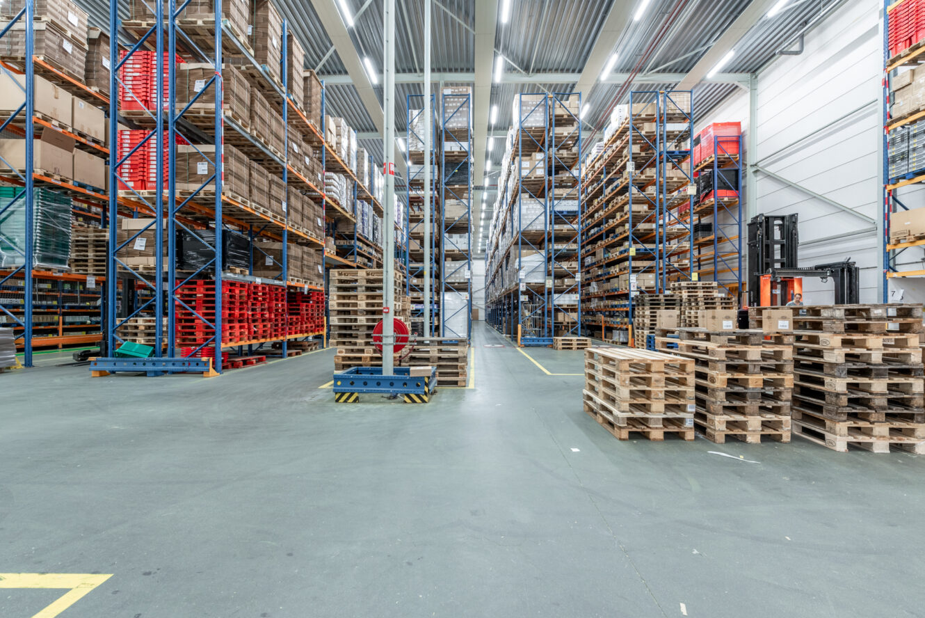 Interieur van een groot magazijn met hoge rekken vol dozen en houten pallets op de vloer.