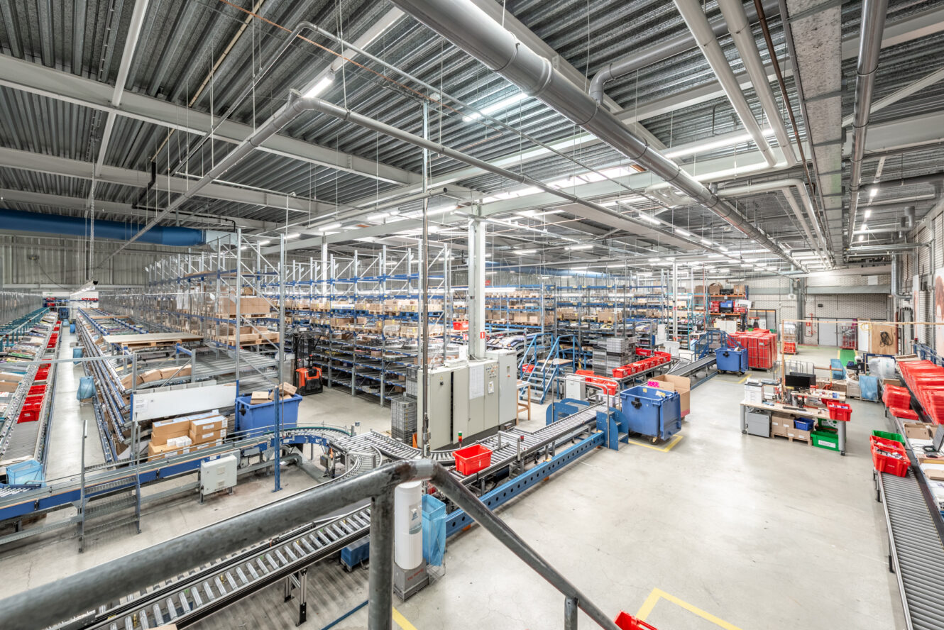 Groot magazijn met metalen rekken, lopende banden en verschillende kratten en dozen die door de ruimte zijn georganiseerd onder heldere industriële verlichting.
