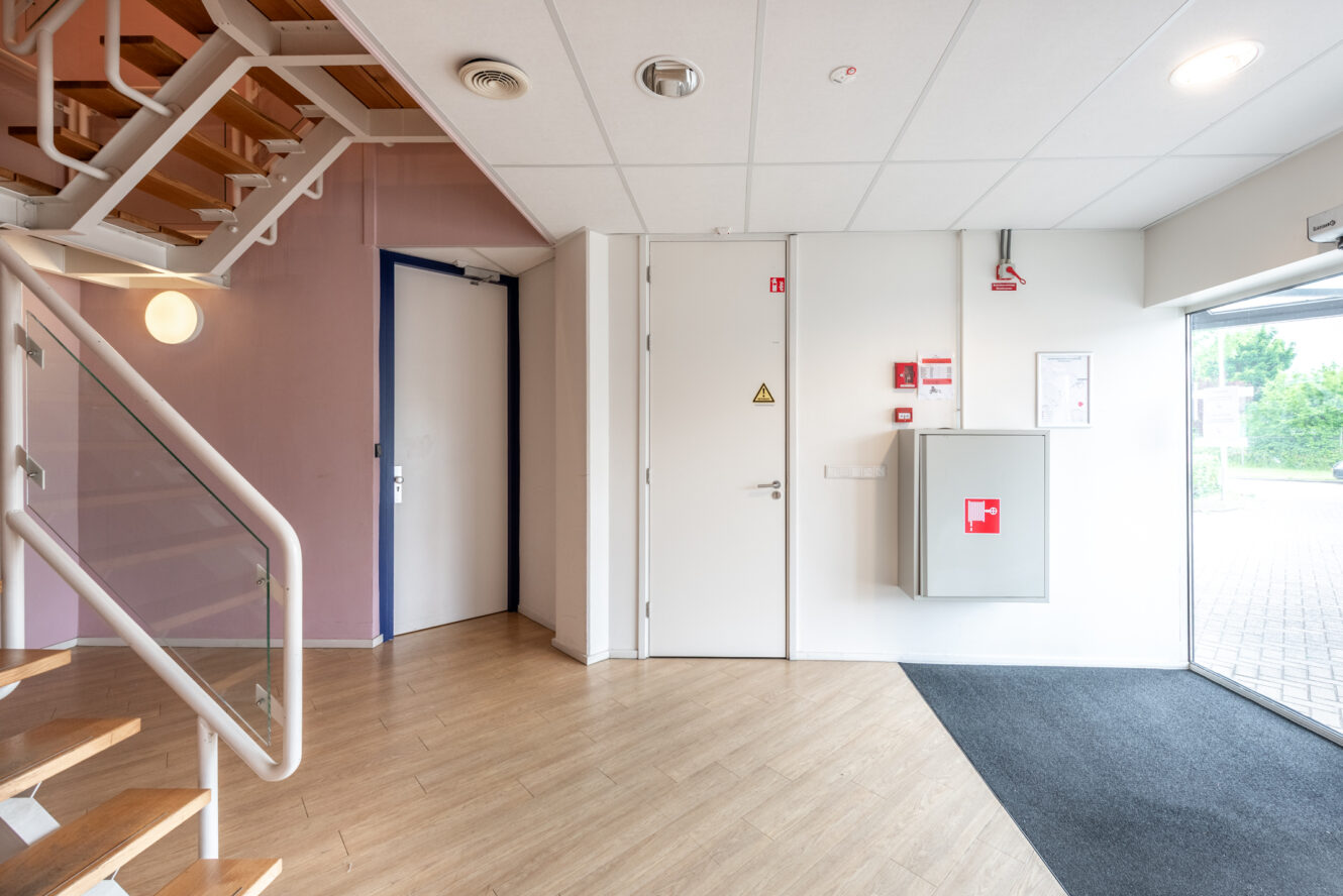 Een schone hal binnenshuis met een trap, witte deuren, een brandveiligheidskast en een groot raam dat natuurlijk licht binnenlaat.