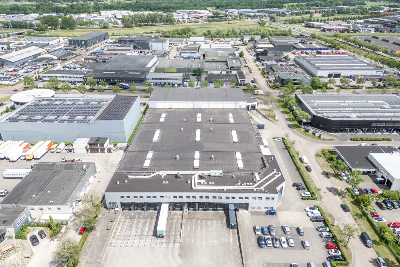 Luchtfoto van een industrieel gebied met grote magazijnen, parkeerplaatsen, vrachtwagens, zonnepanelen en omringend groen.