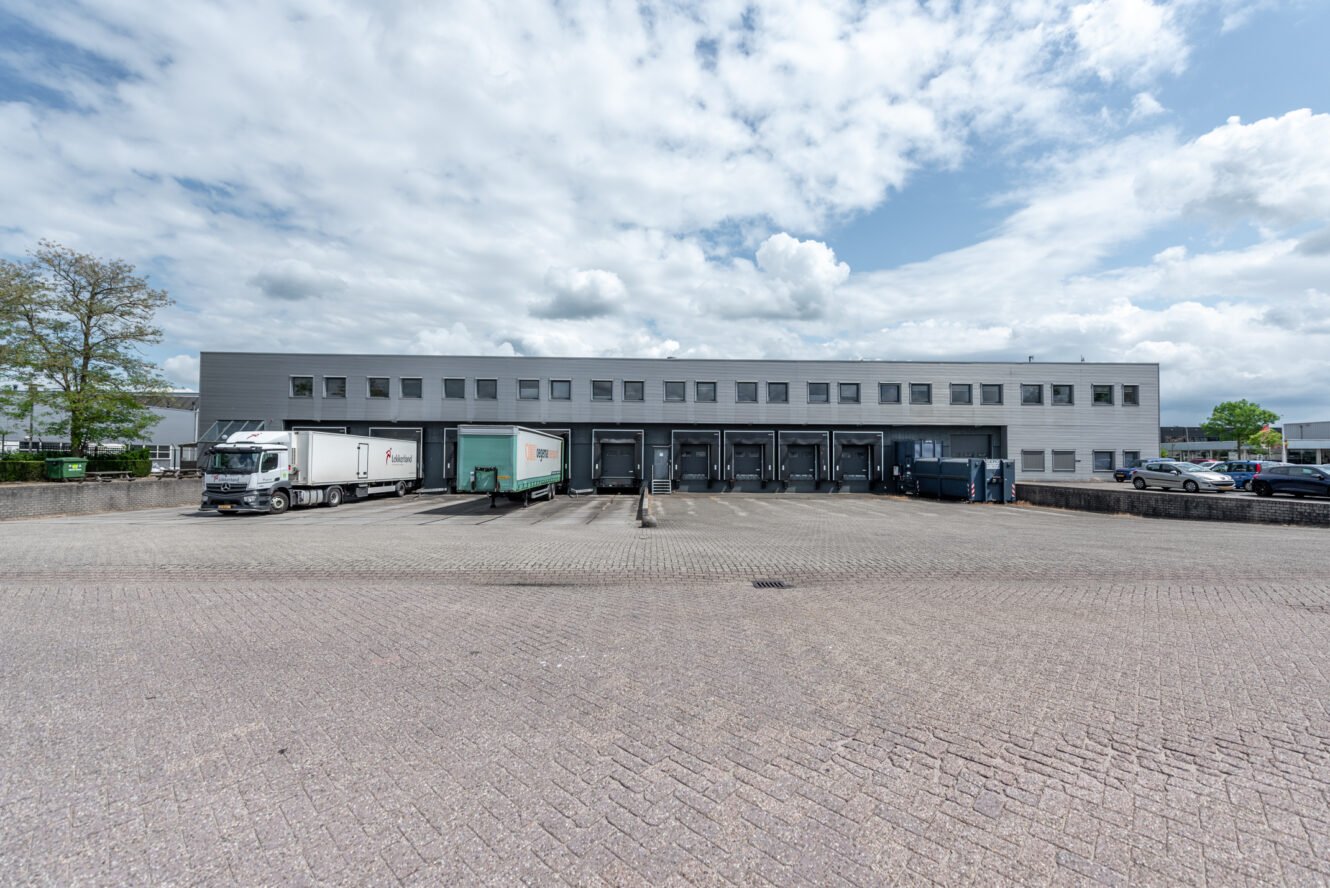 Een groot magazijngebouw met verschillende laadkades, geparkeerde trailers en vrachtwagens onder een gedeeltelijk bewolkte hemel.