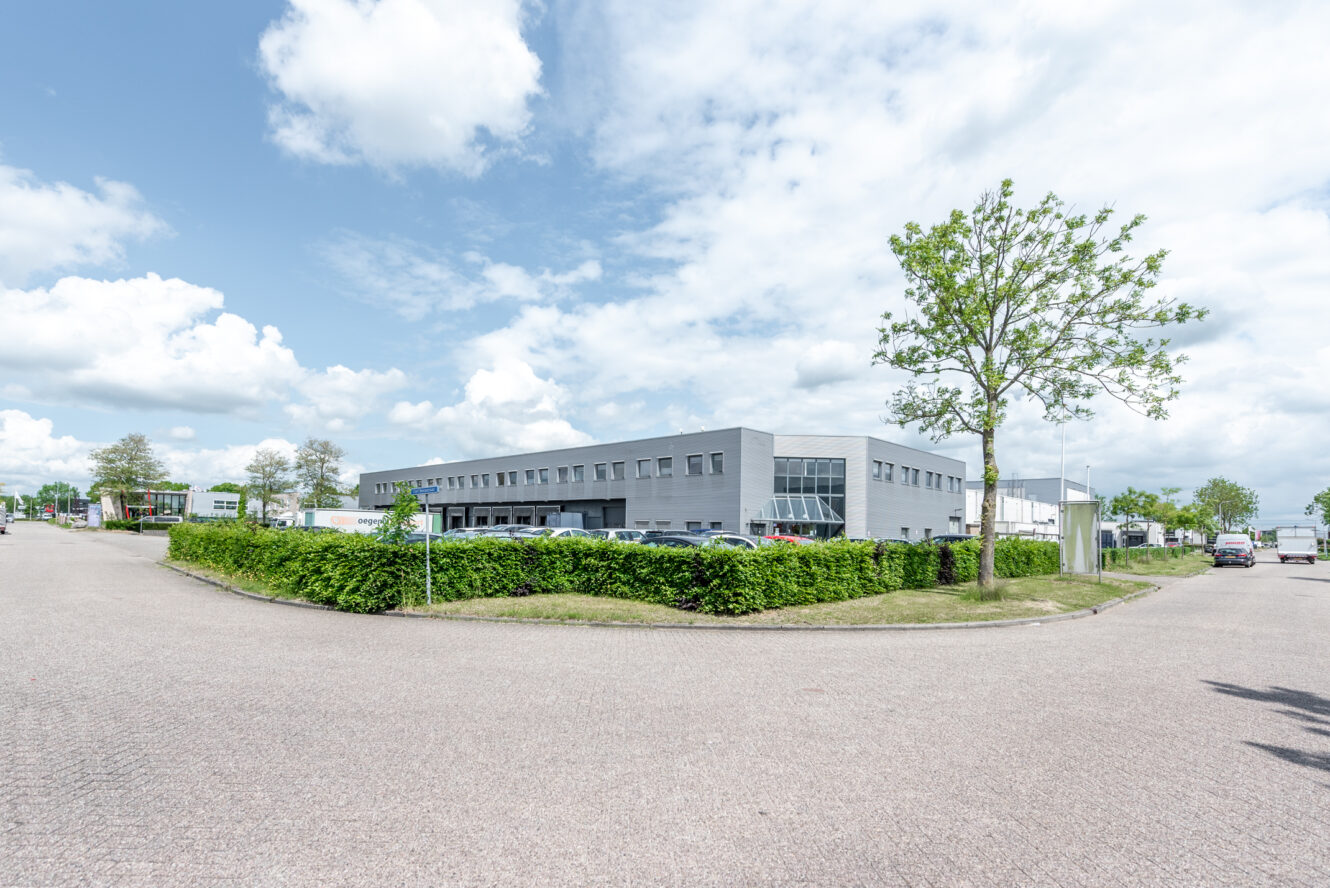 Een modern, grijs industrieel gebouw met grote ramen staat achter een groene haag aan een brede, lege straat onder een gedeeltelijk bewolkte hemel.