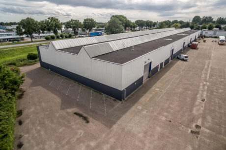 Een groot industrieel pakhuis met een zwart dak en witte muren, omgeven door een leeg geplaveid terrein en wat groen, onder een bewolkte hemel.
