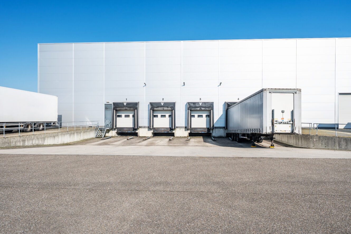 Een wit industrieel magazijn met vier laaddockdeuren; één dock is verbonden met een vrachtwagen, de andere zijn leeg onder een strakblauwe hemel.
