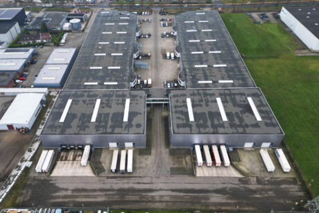 Luchtfoto van een groot industrieel magazijncomplex met meerdere laadkades en trailers die buiten geparkeerd staan. De omgeving bestaat uit grasvelden en aangrenzende gebouwen.