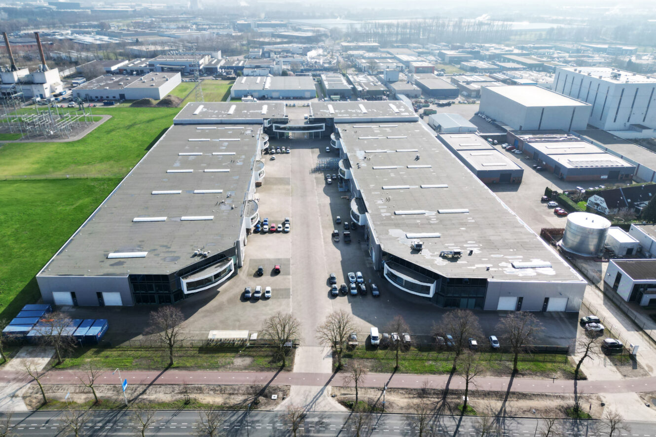 Luchtfoto van twee grote industriële pakhuizen, gescheiden door een centrale parkeerplaats met auto's, omgeven door wegen en groene ruimte.