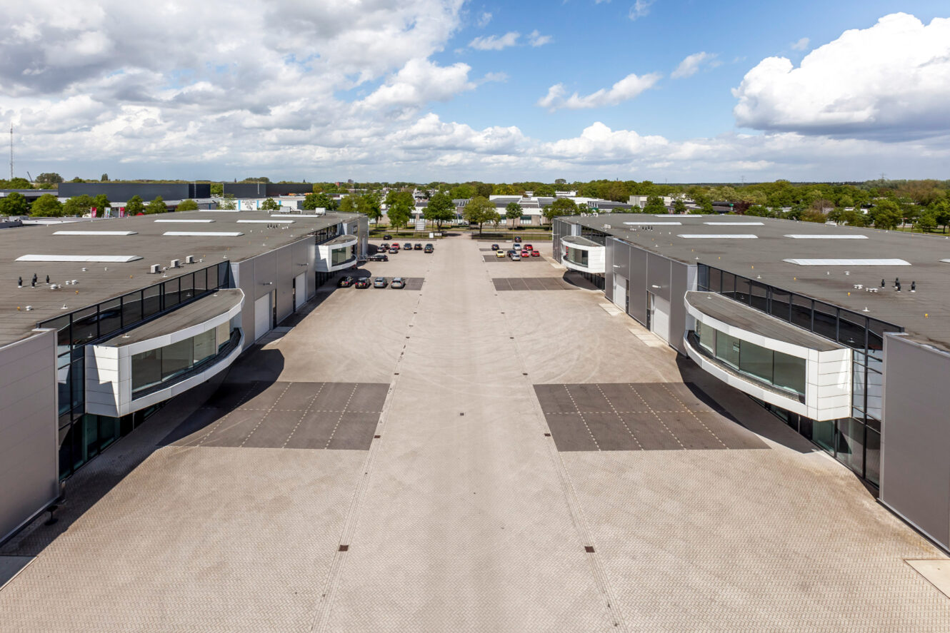 Een breed, leeg industriecomplex met moderne magazijnen aan weerszijden en een verharde centrale oprit onder een gedeeltelijk bewolkte hemel.