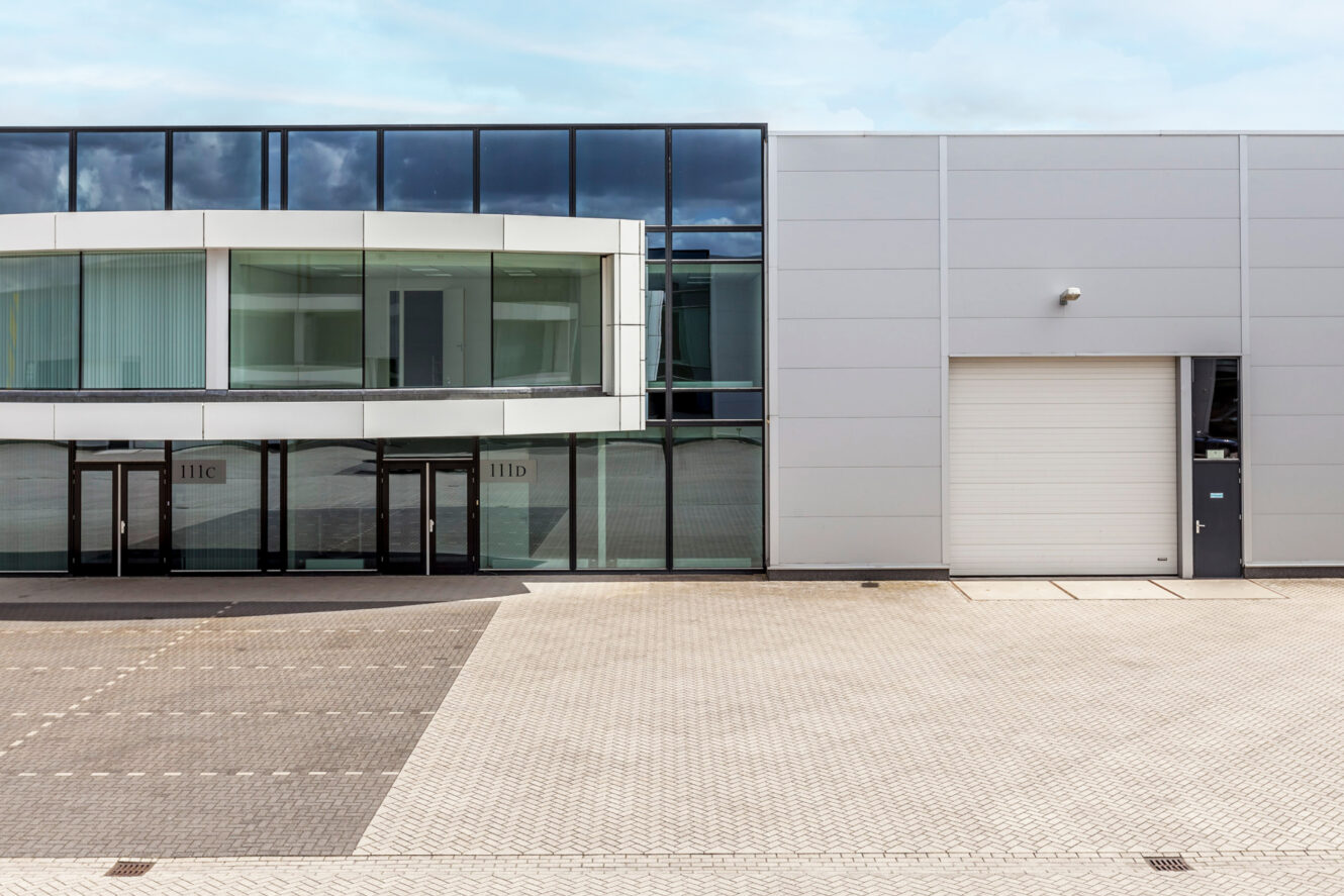 Modern commercieel gebouw aan de buitenkant met grote glazen ramen, een witte paneelgevel, meerdere ingangen en een gesloten roldeur.