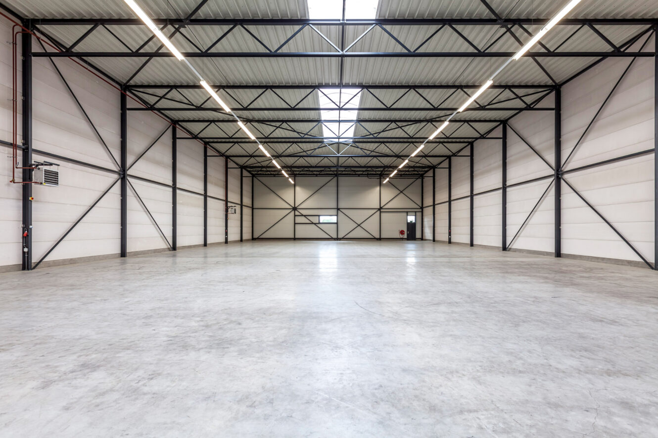 Leeg industrieel pakhuis met een betonnen vloer, witte muren, zichtbare stalen balken en overheadverlichting. Er zijn een paar ramen en een deur in de achterwand.