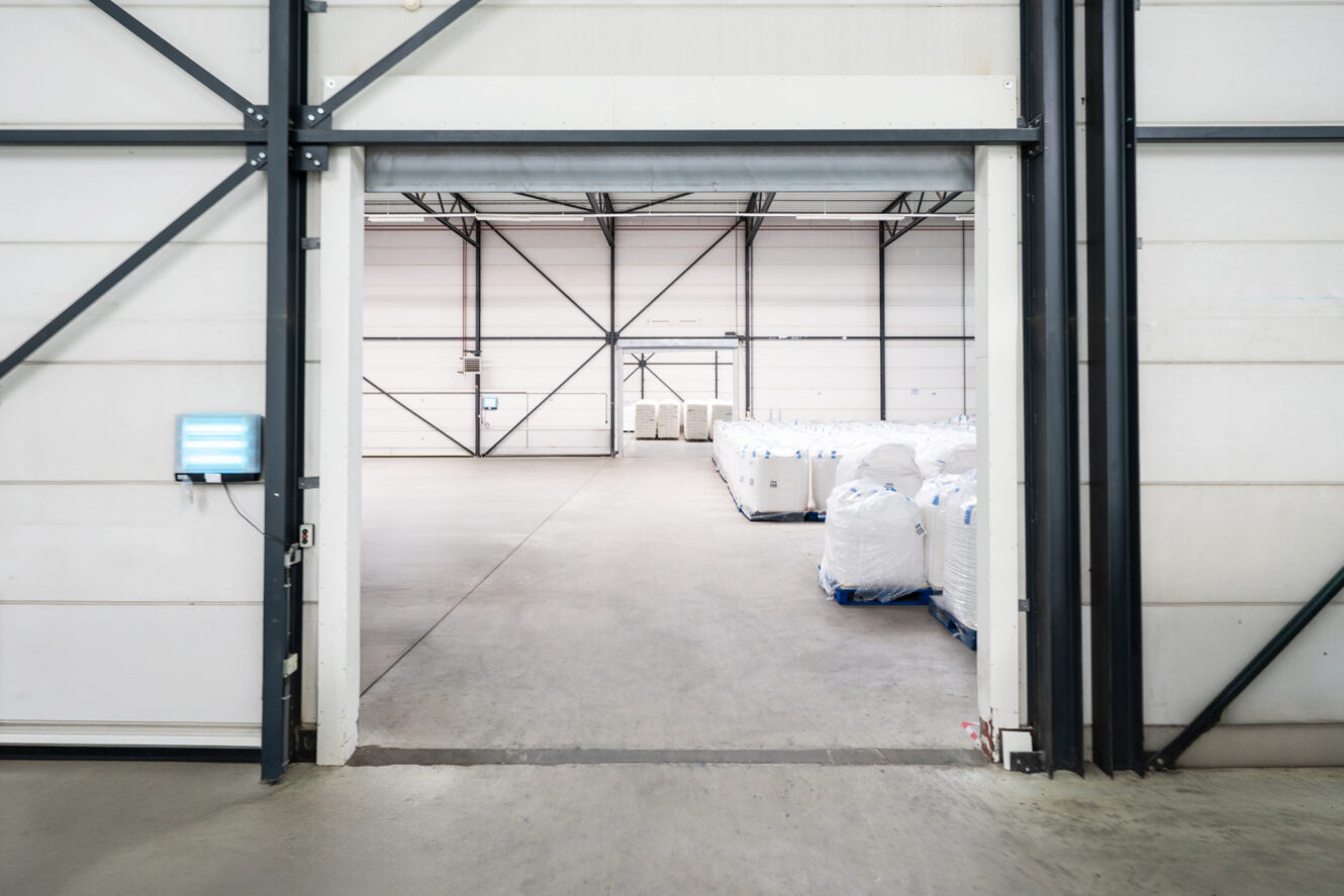Een groot industrieel magazijn met witte muren, zichtbare stalen balken en gestapelde witte zakken op pallets binnenin.