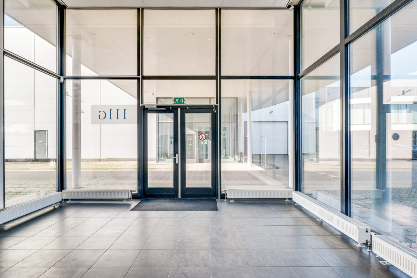 Een moderne glazen ingang met dubbele deuren, tegelvloer en grote ramen. Het woord 111C staat omgekeerd op het glas. Buiten zijn gebouwen zichtbaar.