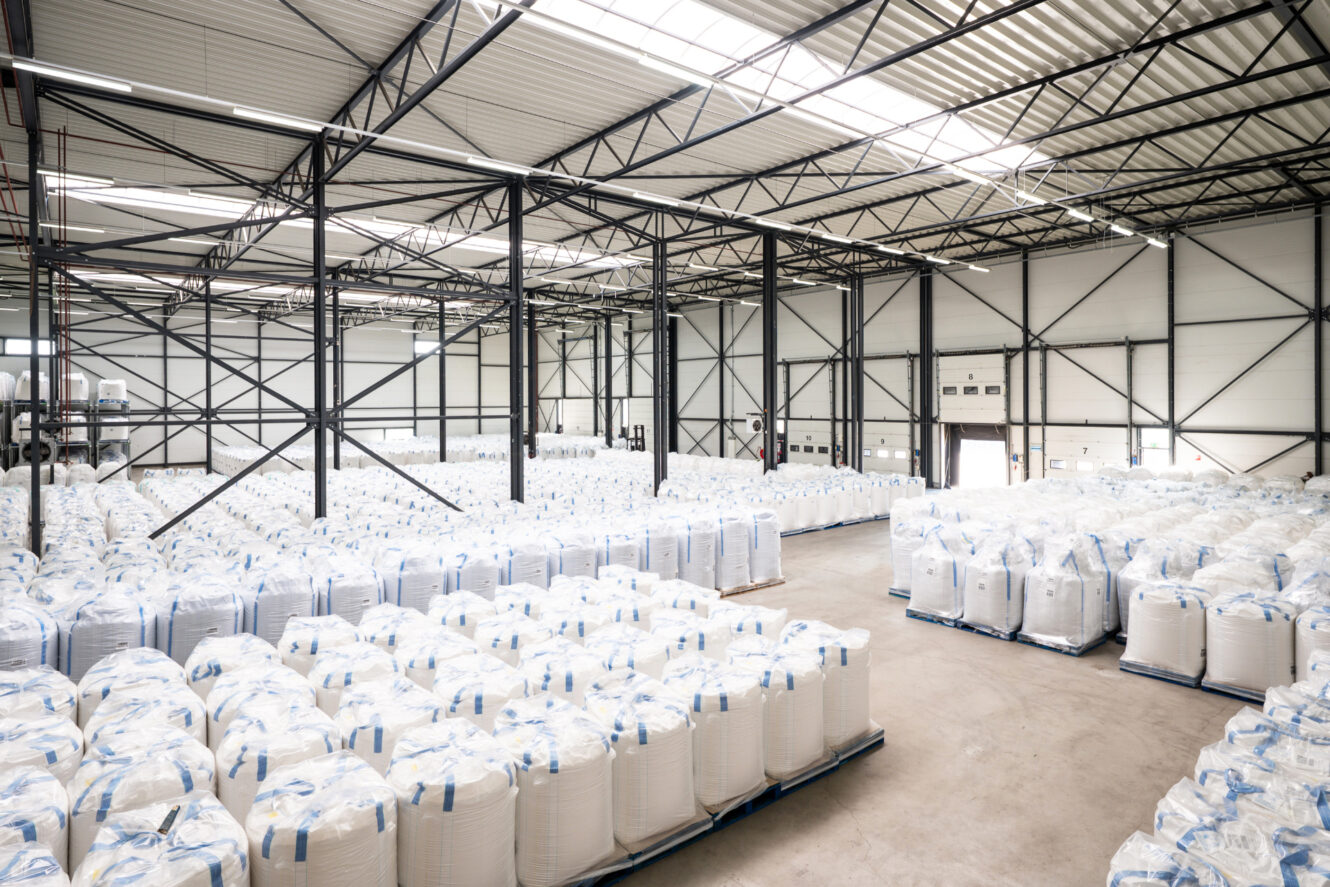 Groot interieur van magazijn gevuld met netjes gestapelde witte industriële zakken op pallets, opgesteld in rijen onder een metalen dak met dakramen.