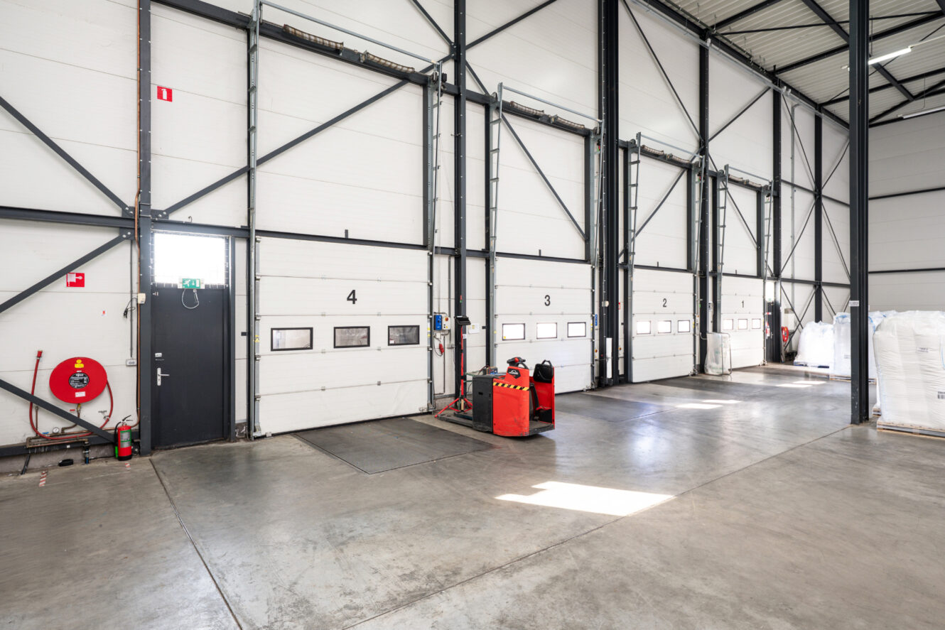 Een magazijninterieur met vijf genummerde laadperrondeuren, palletvijzels, brandbeveiligingsapparatuur en betonnen vloeren onder heldere verlichting.
