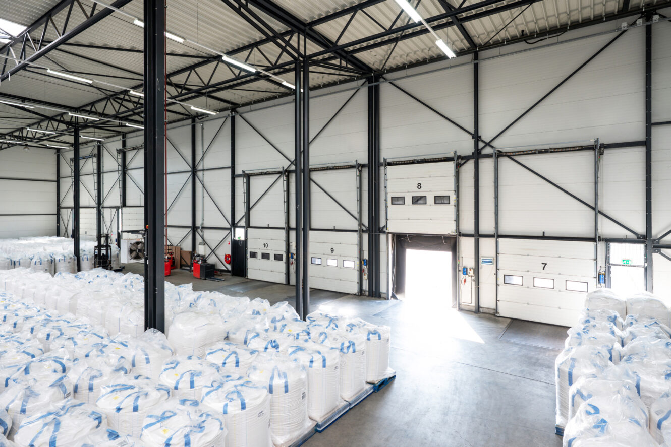 Groot magazijninterieur met hoge plafonds, gevuld met witte bulkzakken op pallets; verschillende laaddockdeuren, waarvan één open, die daglicht binnenlaten.