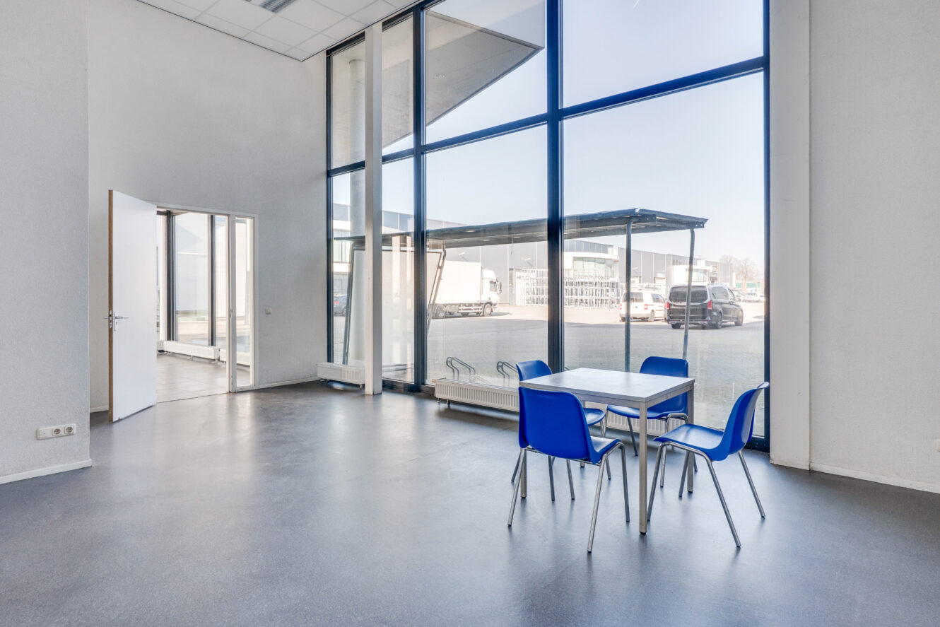 Een kleine vierkante tafel met vier blauwe stoelen staat bij grote ramen van vloer tot plafond in een lichte, lege kamer met uitzicht op geparkeerde voertuigen buiten.