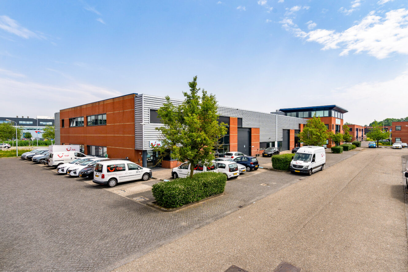 Een commercieel bedrijventerrein met moderne bakstenen en metalen gebouwen, meerdere geparkeerde auto's en een heldere hemel.