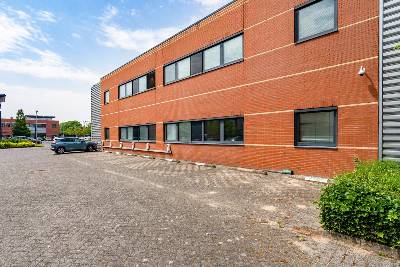Een bakstenen kantoorgebouw met grote ramen en een aangrenzend parkeerterrein met een enkele geparkeerde auto, onder een gedeeltelijk bewolkte hemel.