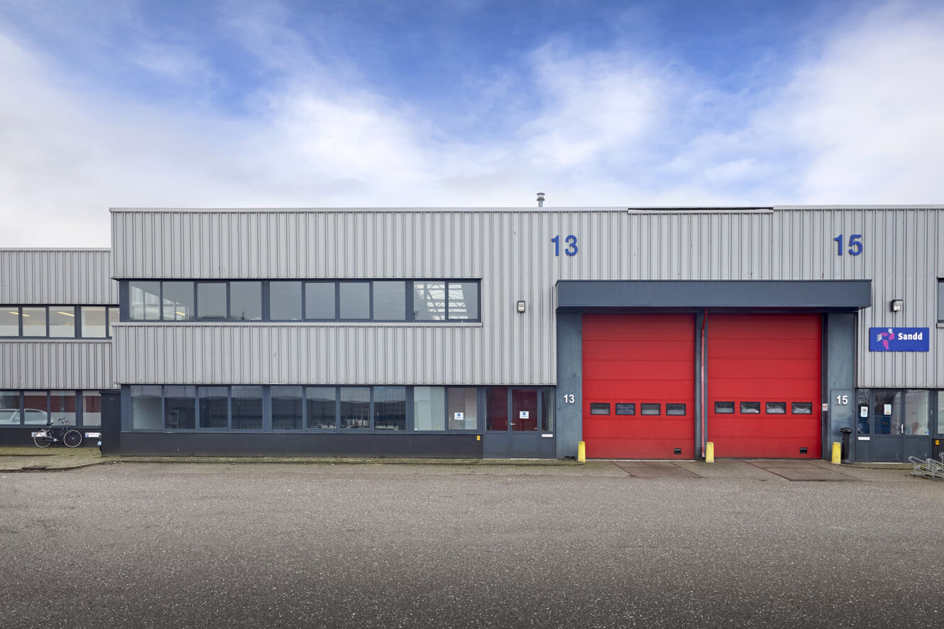 Industrieel pakhuis met grijze metalen bekleding, grote rode garagedeuren genummerd 13 en 15 en een rij ramen op de eerste verdieping.