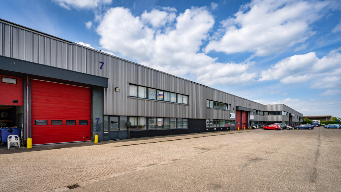 Een rij moderne industriële magazijnen met grijze metalen wanden, rode laaddeuren en geparkeerde voertuigen onder een gedeeltelijk bewolkte hemel.