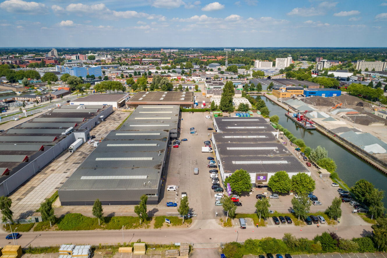 Luchtfoto van een industriegebied met grote magazijnen, parkeerplaatsen, auto's, een kanaal en stadsgebouwen op de achtergrond onder een gedeeltelijk bewolkte hemel.