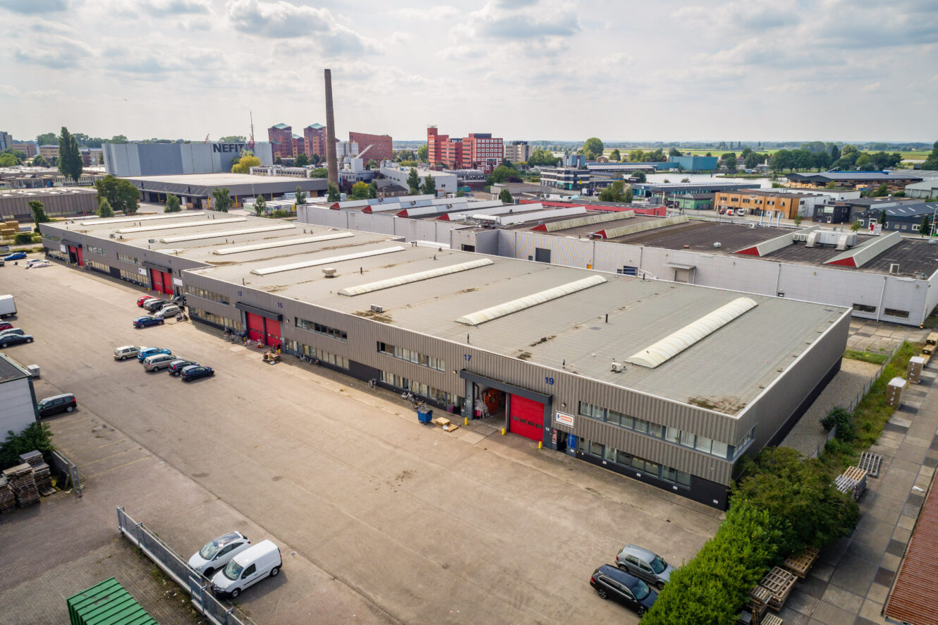 Luchtfoto van een groot industrieel magazijncomplex met meerdere rode deuren, geparkeerde auto's en omliggende gebouwen onder een gedeeltelijk bewolkte hemel.