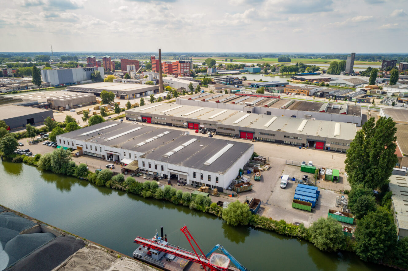 Luchtfoto van een industriegebied met grote magazijnen, fabrieken, waterweg op de voorgrond en woongebouwen op de achtergrond onder een bewolkte hemel.