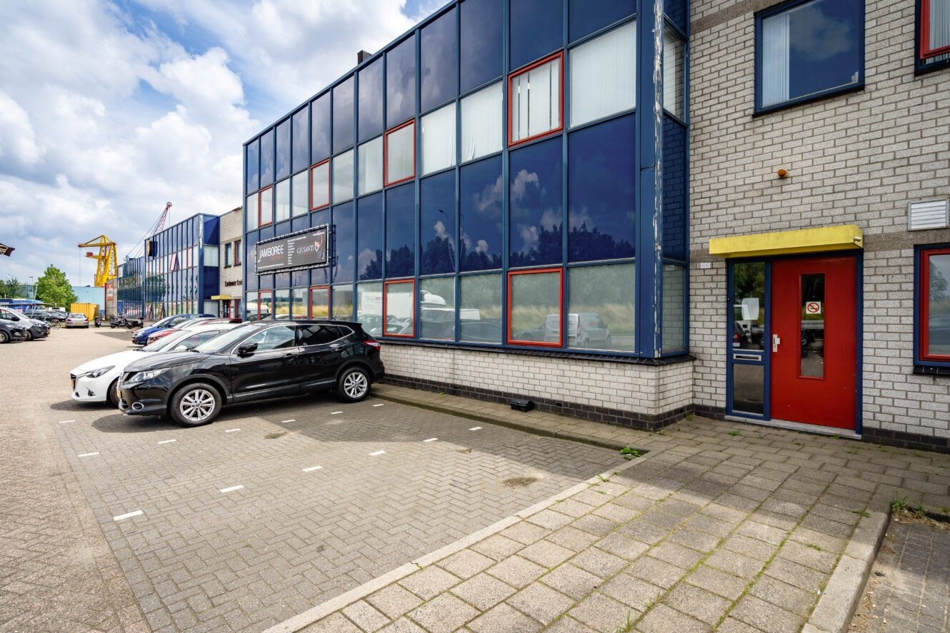 Een modern industrieel gebouw met blauwgetinte ramen, een aantal geparkeerde auto's en een rode toegangsdeur onder een gele luifel.