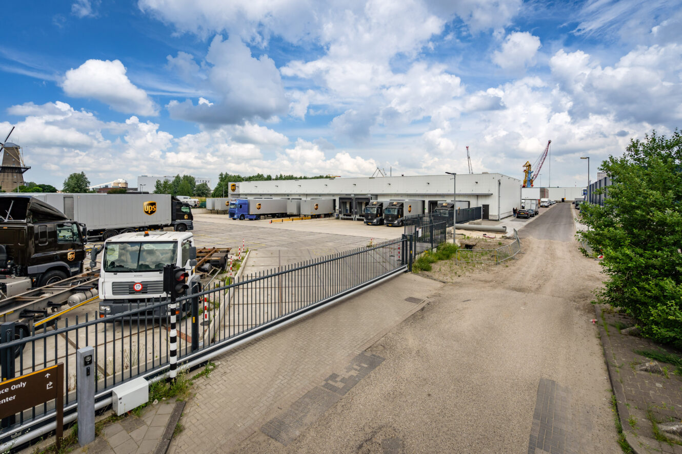 Een omheind industrieterrein met geparkeerde UPS bestelwagens en trailers naast een groot wit magazijn onder een gedeeltelijk bewolkte hemel.