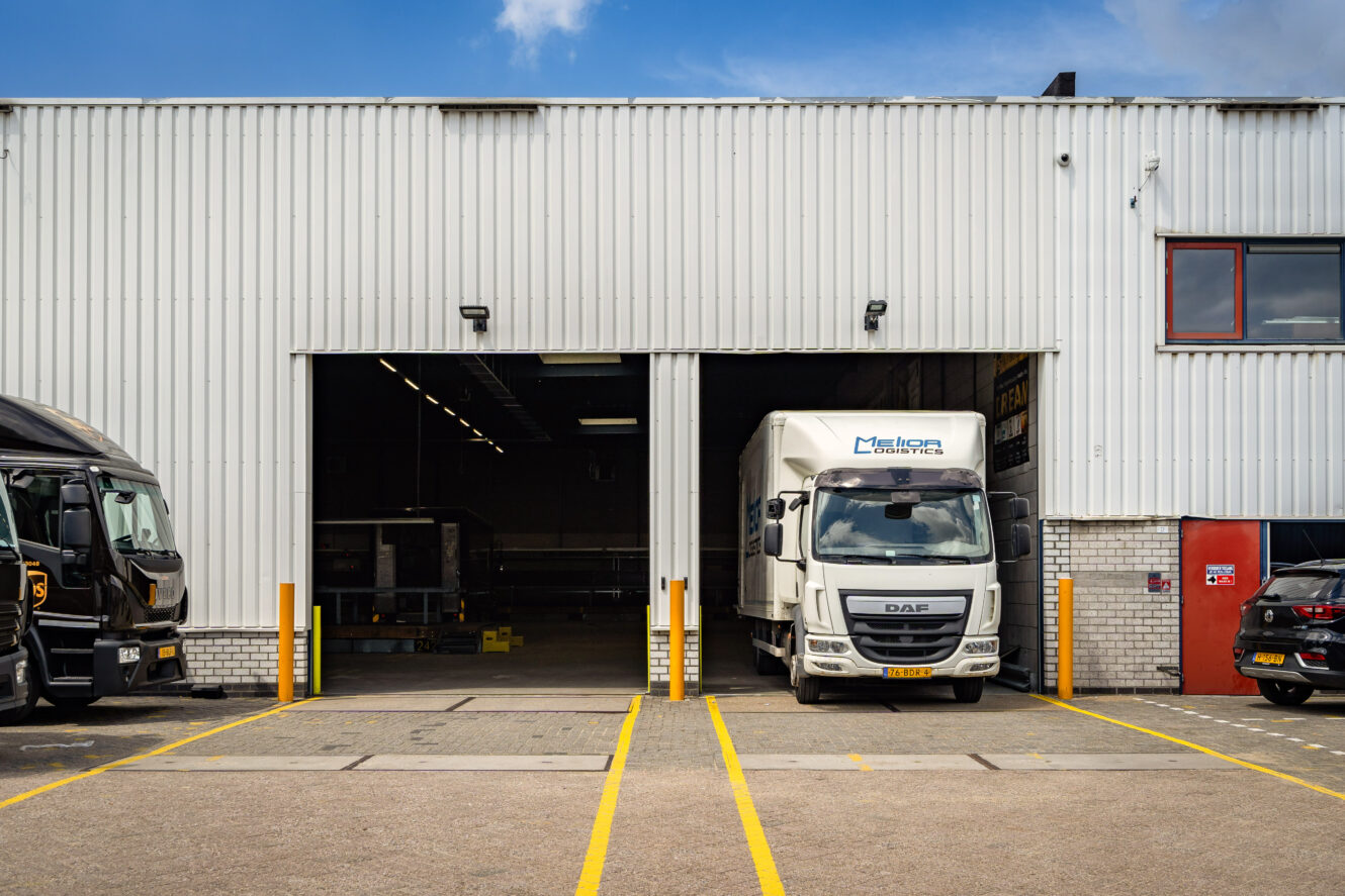 Een witte bestelwagen staat geparkeerd voor een grote magazijningang, met een andere donkere vrachtwagen aan de linkerkant en gele lijnen die parkeerplaatsen markeren op de grond.