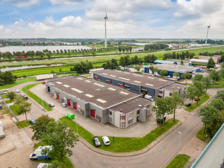 Luchtfoto van een industrieel complex met meerdere magazijngebouwen, nabij geparkeerde auto's, wegen en windturbines in een groen landschap onder een gedeeltelijk bewolkte hemel.