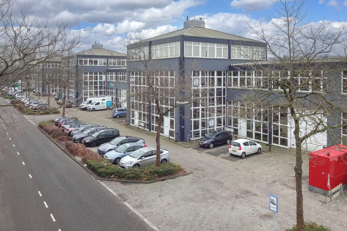 Een rij geparkeerde auto's voor een groot, modern industrieel gebouw met veel ramen, bladerloze bomen en een grotendeels lege weg onder een gedeeltelijk bewolkte hemel.