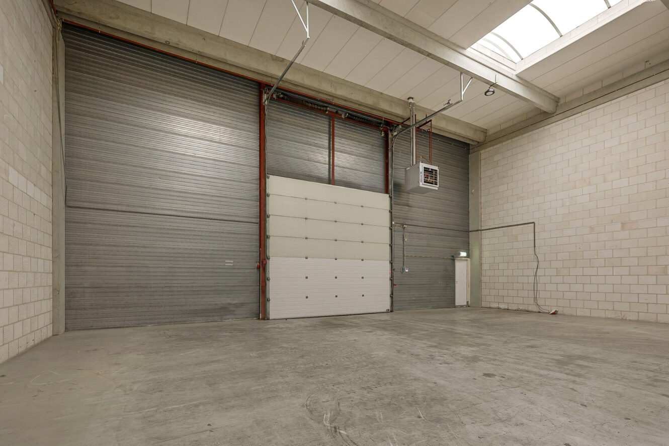 Leeg industrieel magazijninterieur met betonnen vloer, hoog plafond, grote roldeuren en witte bakstenen muren. Natuurlijk licht valt binnen via dakramen in het dak.