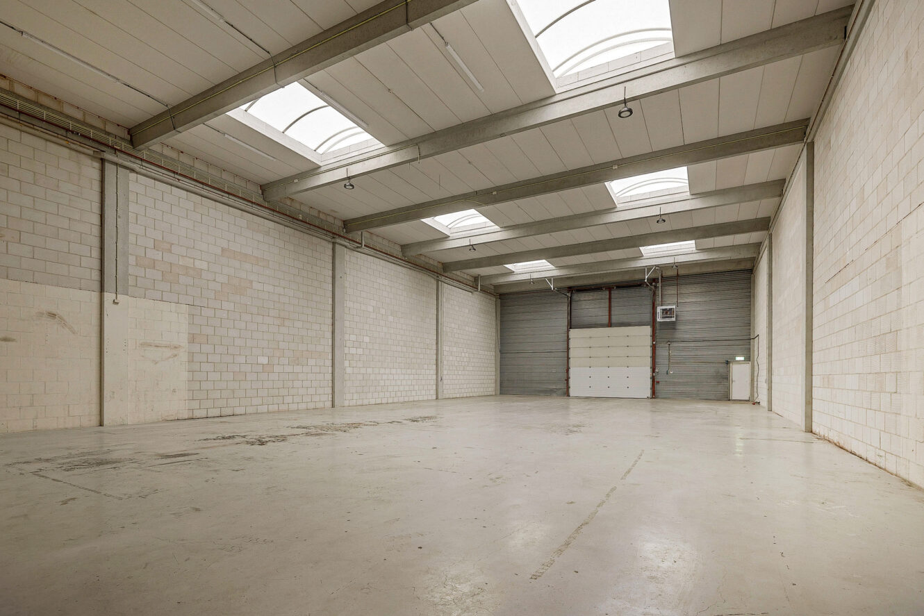 Leeg pakhuis met witte bakstenen muren, betonnen vloer, hoog plafond, dakramen en grote gesloten roldeuren aan het uiteinde.