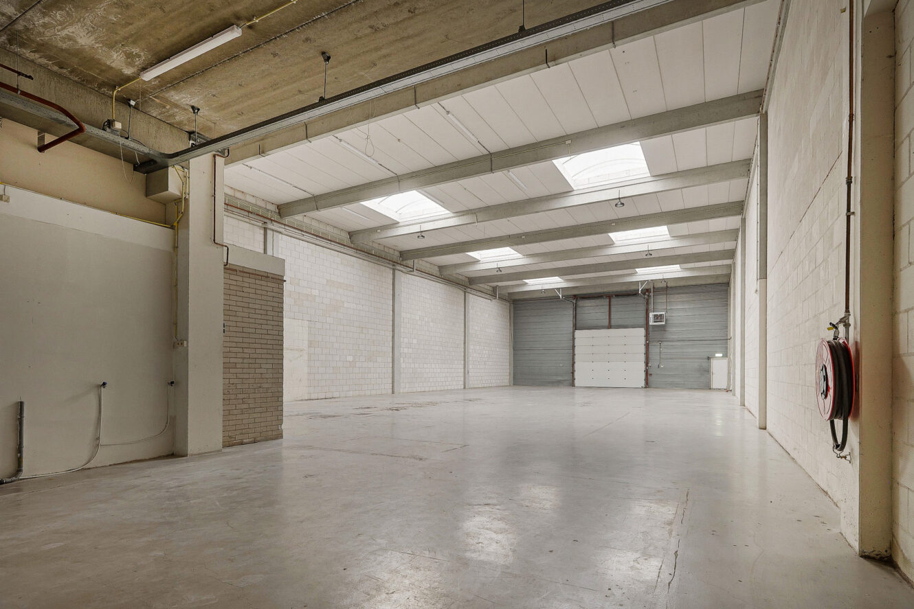 Groot leeg pakhuis met betonnen vloer, hoog plafond, dakramen, witte bakstenen muren en een gesloten garagedeur aan het uiteinde. Aan de rechtermuur is een brandslanghaspel gemonteerd.