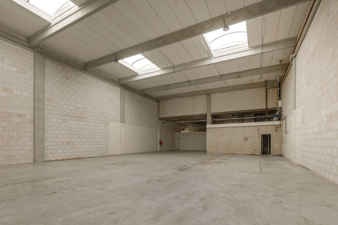 Ruim, leegstaand industrieel pakhuis met hoge plafonds, witte bakstenen muren, betonnen vloer en dakramen die natuurlijk licht binnenlaten.