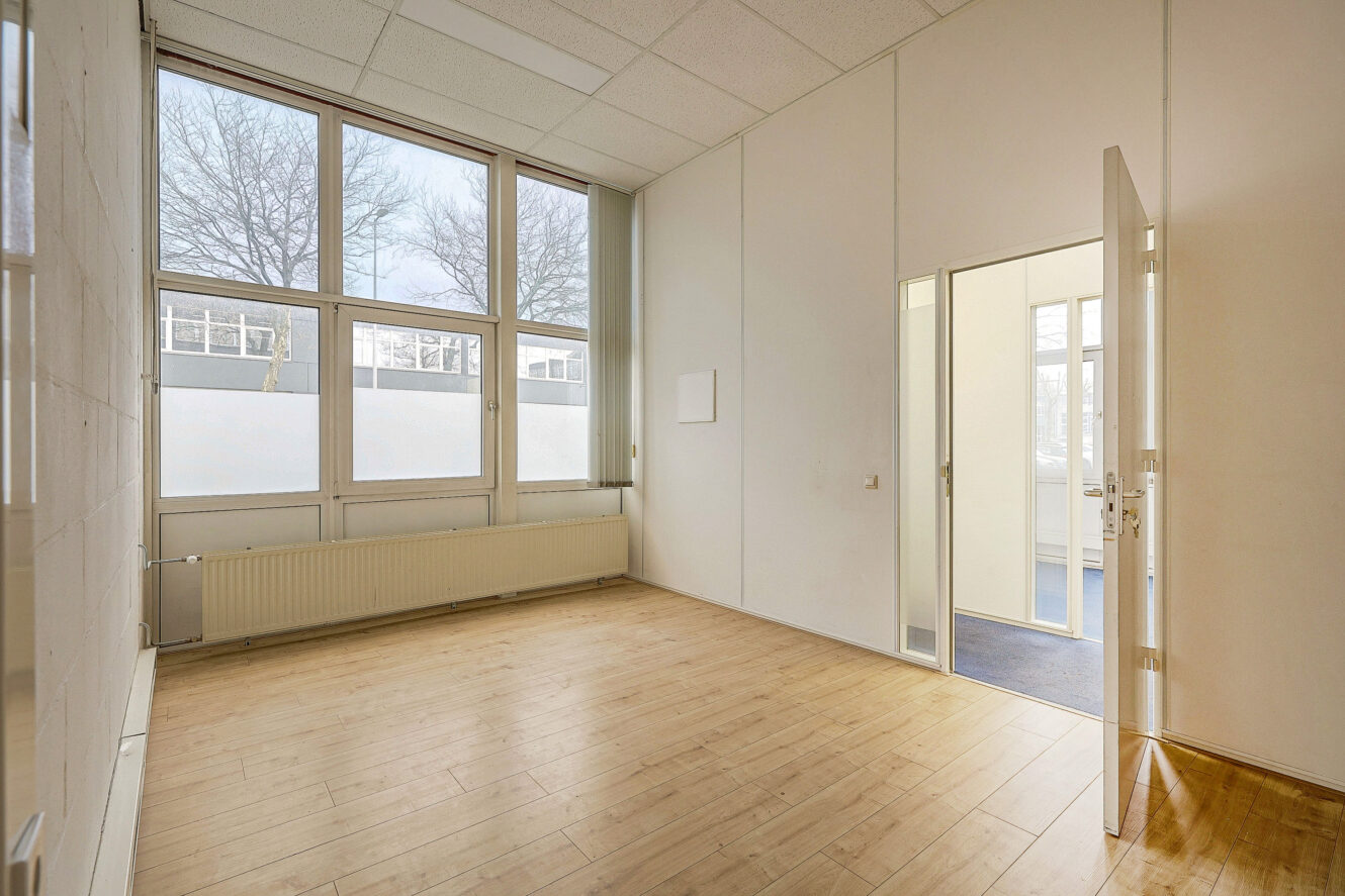 Lege kamer met lichte houten vloer, grote ramen, witte muren, een radiator onder de ramen en een open deur die leidt naar een aangrenzende ruimte met blauw tapijt.