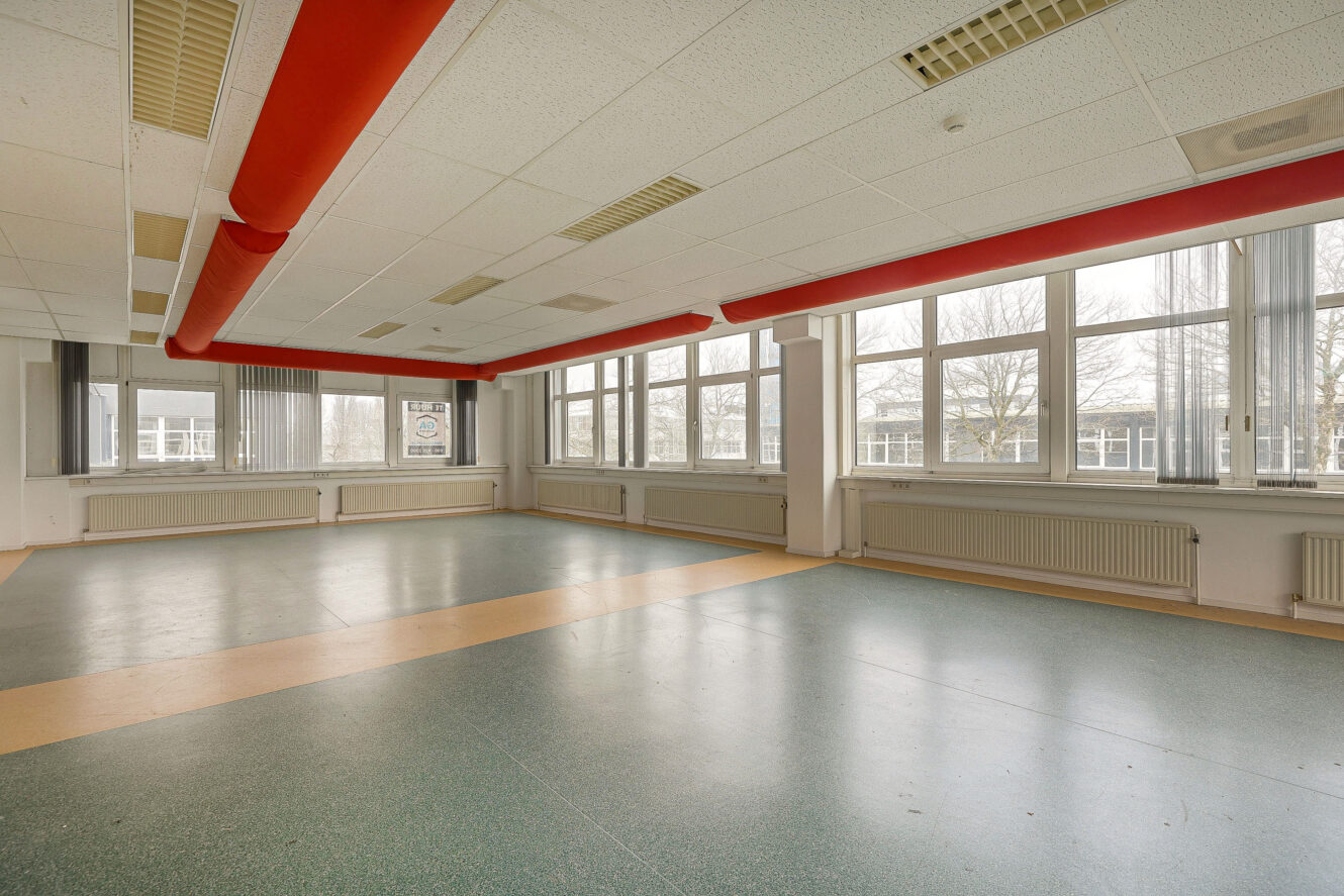 Grote, lege kamer met groene linoleumvloer, witte muren, grote ramen langs twee kanten, rode zichtbare plafondpijpen en tl-verlichting.