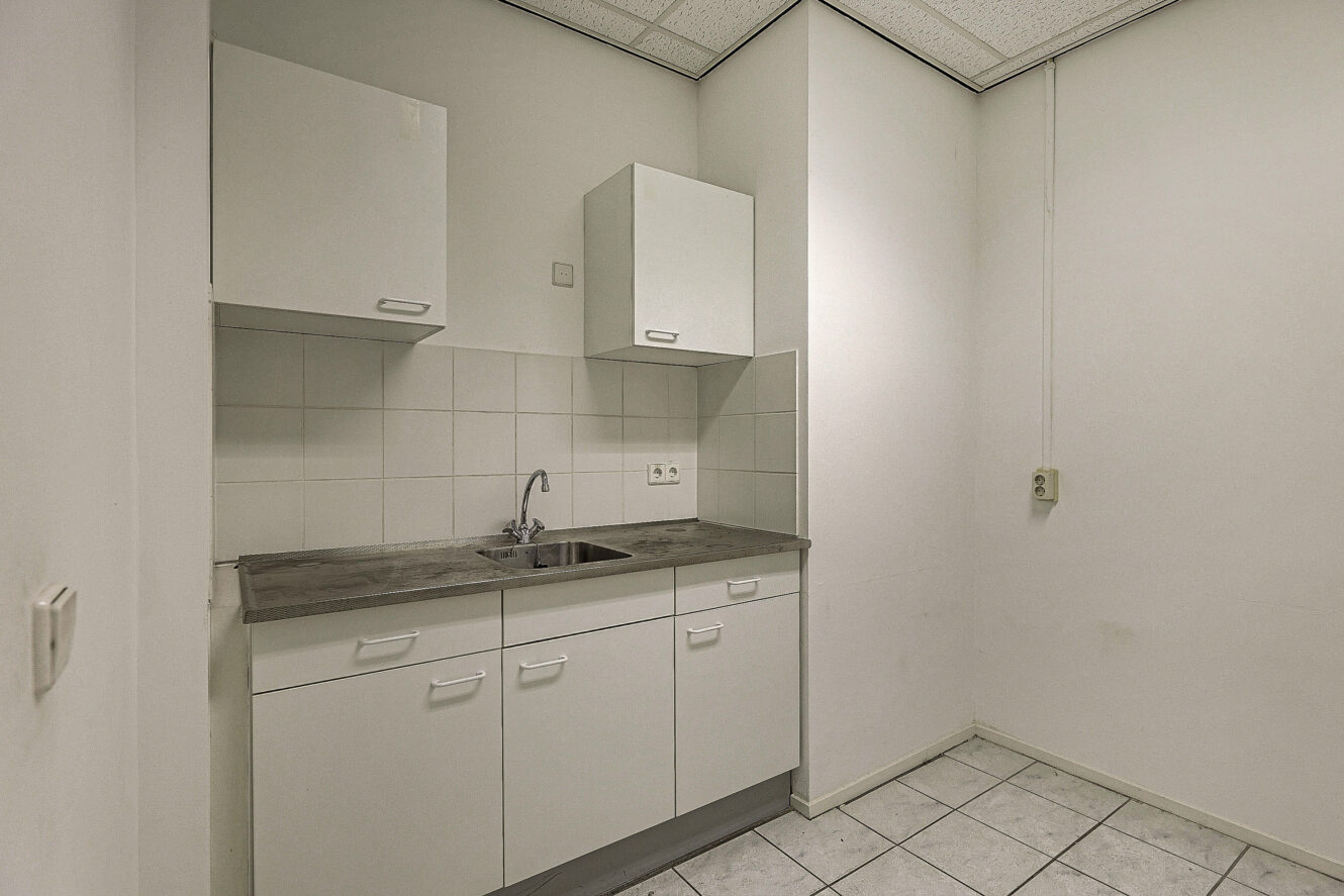 Kleine keuken met witte kastjes, een enkele gootsteen, betegelde achterwand en betegelde vloer. De muren zijn effen en er is een zichtbaar stopcontact aan de rechterkant.