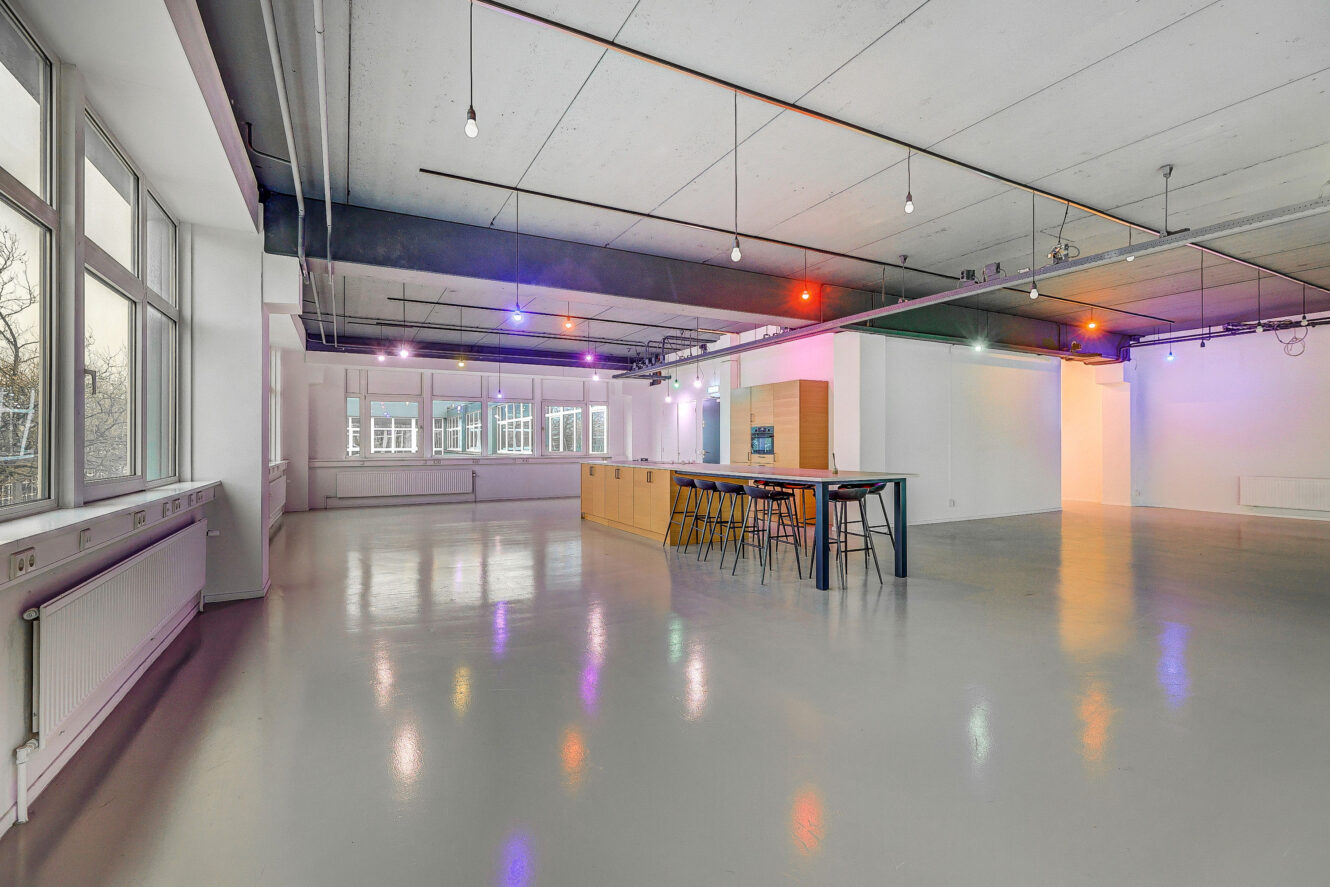 Ruime moderne kamer met gepolijste vloeren, grote ramen, een zichtbaar plafond, een lange houten tafel met krukken en minimale verlichting.