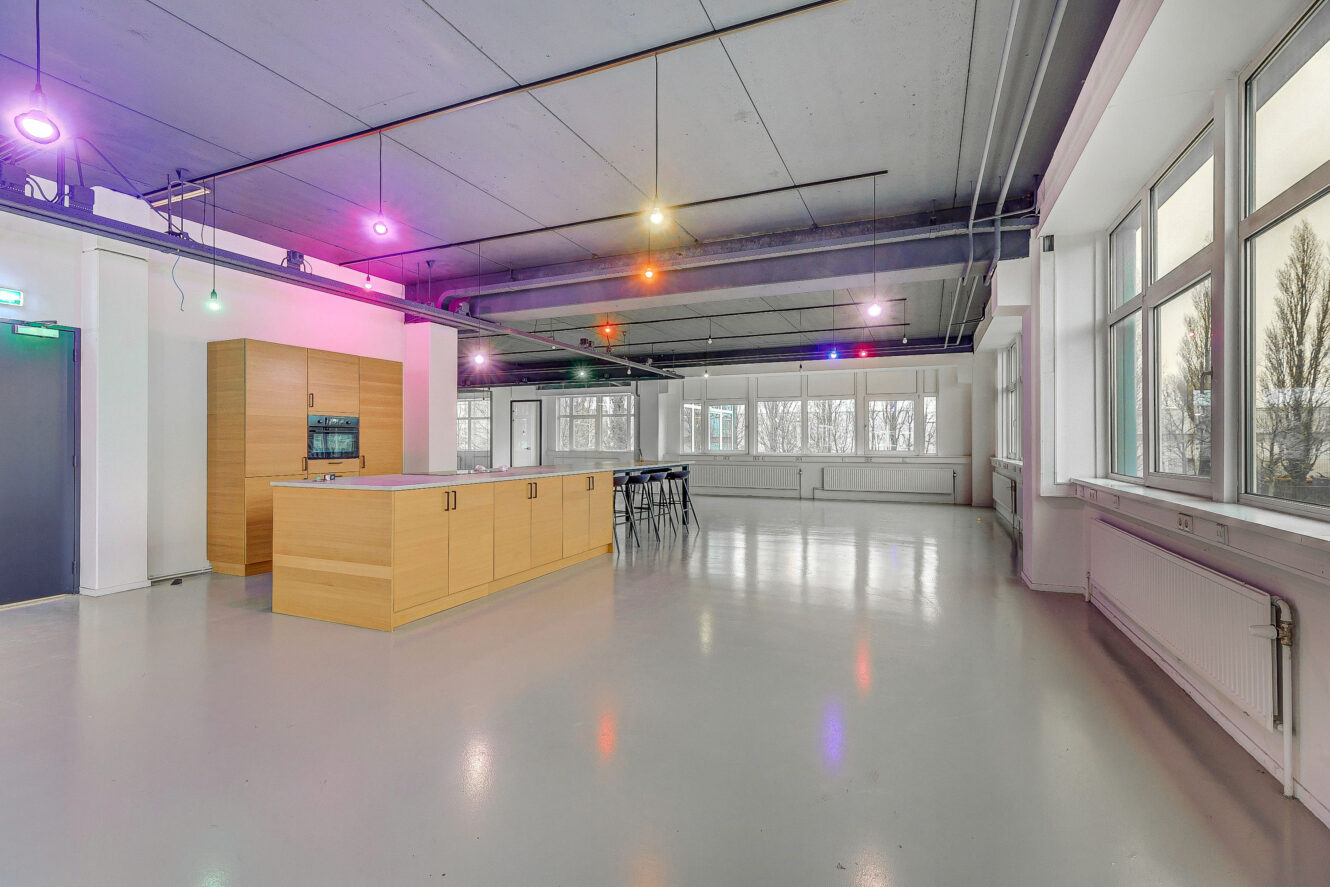 Ruime moderne keuken met een houten eiland, hoge krukken, grote ramen, gepolijste vloer, blootgesteld plafond en kleurrijke plafondverlichting.